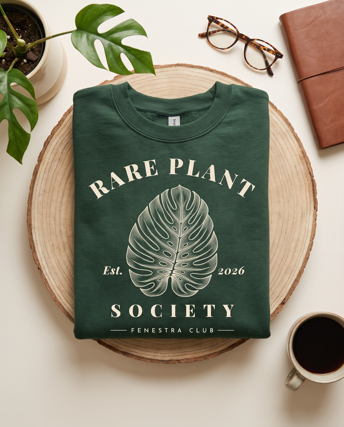 Rare Plant Society sweatshirt in Forest Green folded on a wooden board with coffee and a monstera leaf