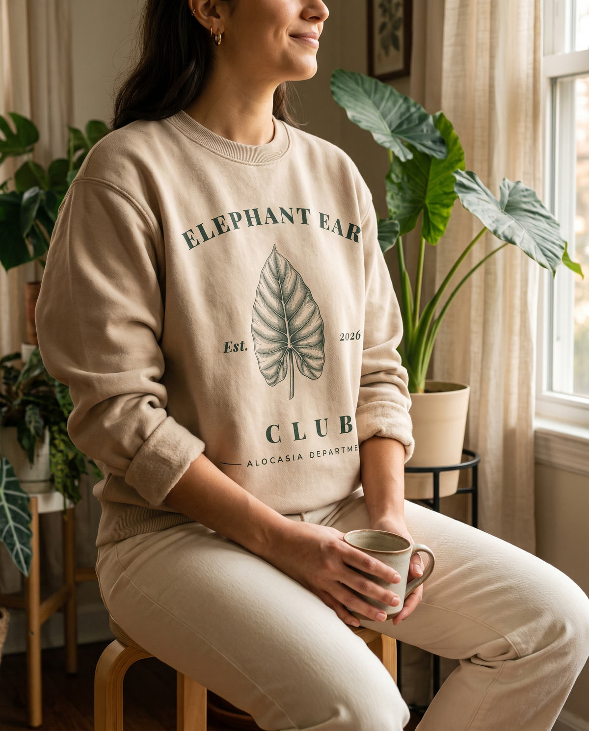 Woman with a mug in a plant-filled living room, wearing the Elephant Ear Club crewneck in Sand