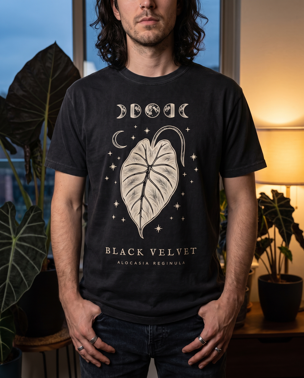 Man in a moody plant-filled apartment wearing the Black Velvet alocasia tee in Black
