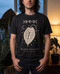 Man in a moody plant-filled apartment wearing the Black Velvet alocasia tee in Black